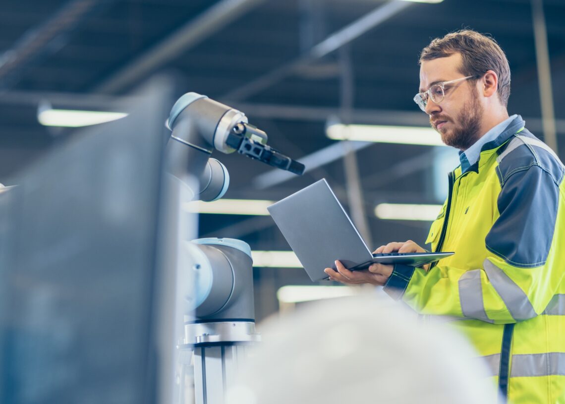 A factory employee wears a yellow safety vest as he works on a laptop in front of a robotic machine.