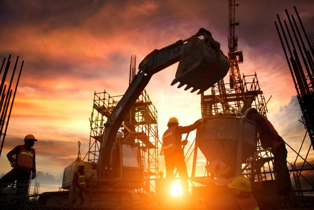 A team of construction professionals wearing hard hats surround a concrete mixer. Behind them is an excavator.