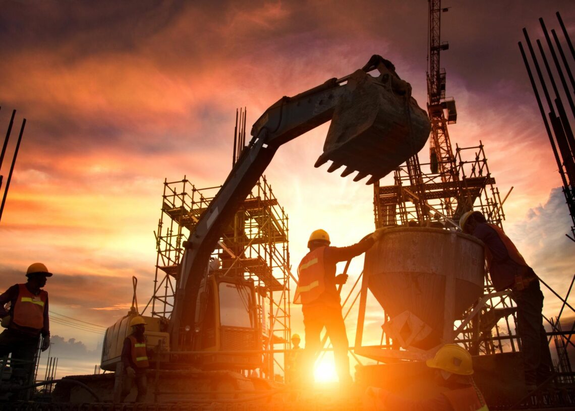 A team of construction professionals wearing hard hats surround a concrete mixer. Behind them is an excavator.