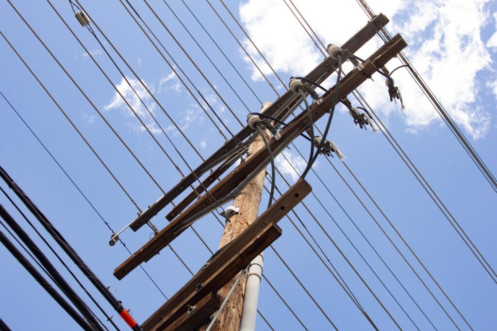 A utility pole with power lines running across it. Behind the pole is a blue sky with a few white clouds.