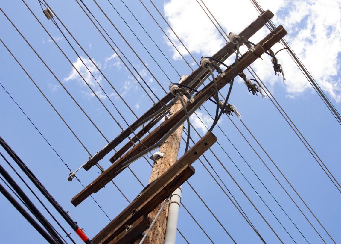 A utility pole with power lines running across it. Behind the pole is a blue sky with a few white clouds.