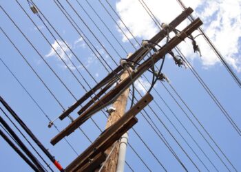 A utility pole with power lines running across it. Behind the pole is a blue sky with a few white clouds.