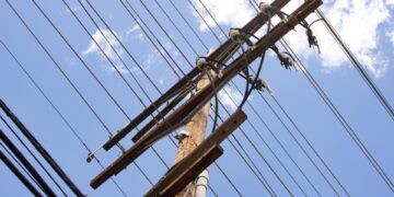 A utility pole with power lines running across it. Behind the pole is a blue sky with a few white clouds.