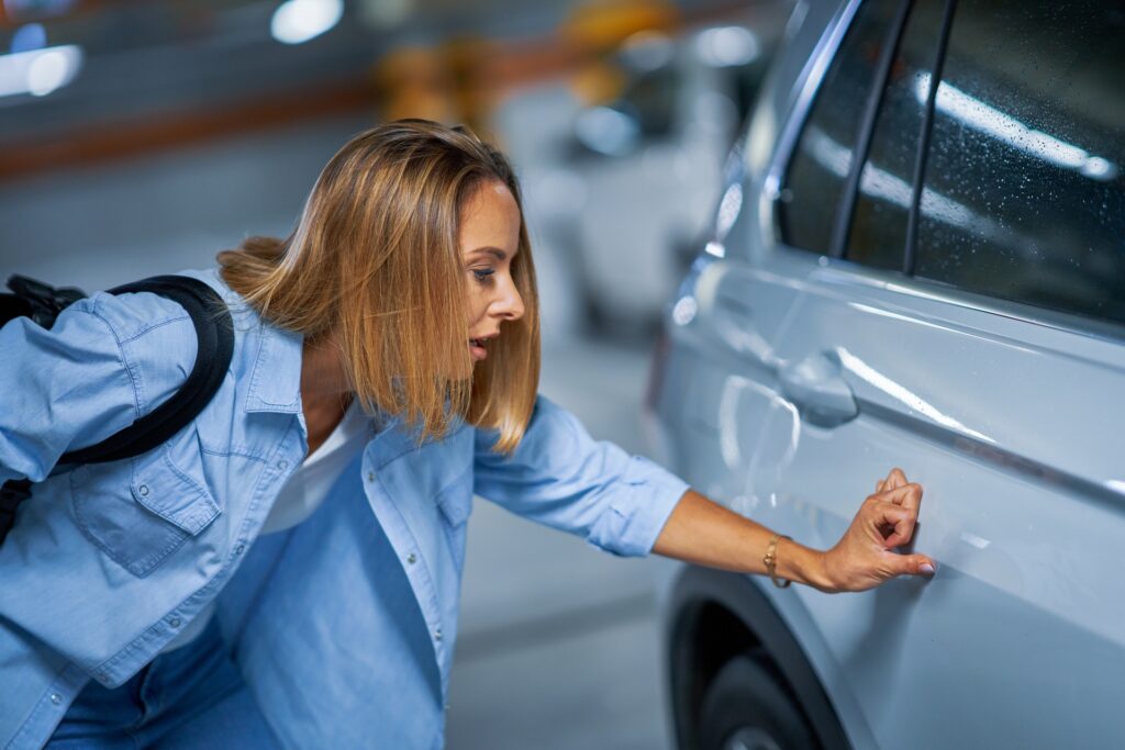 A young woman looking down at a scratch on her back car door. She is running her thumb over the scratch.