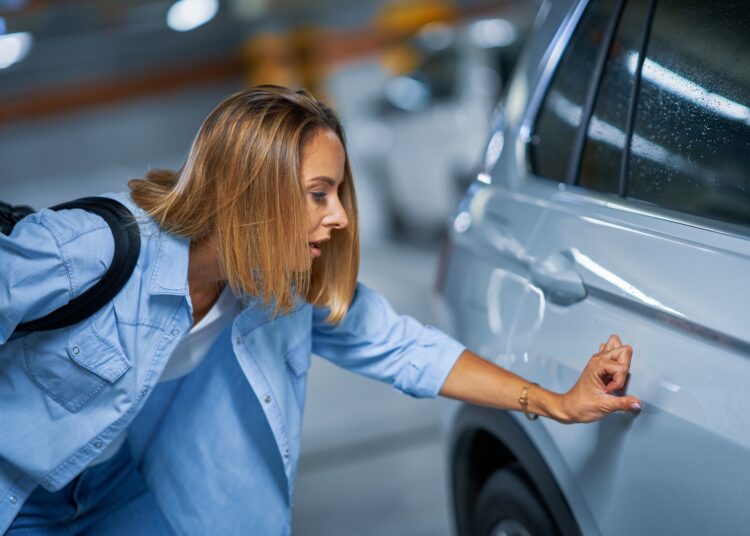 A young woman looking down at a scratch on her back car door. She is running her thumb over the scratch.