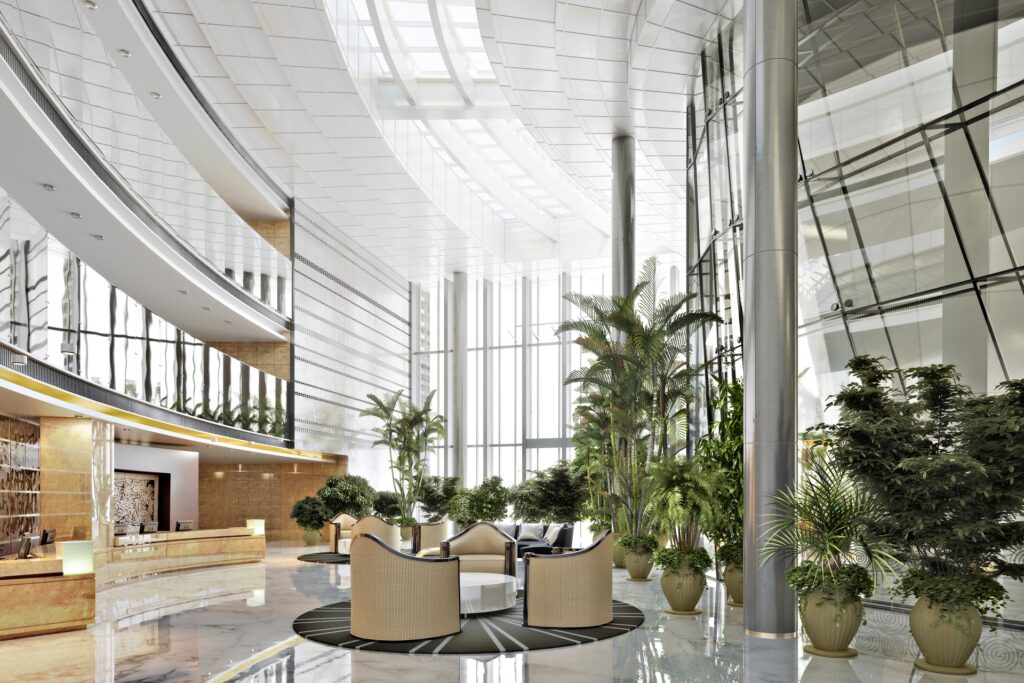 A building with a large lobby. There are ceiling-high windows in the back ground and greenery all around.
