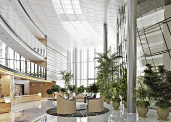 A building with a large lobby. There are ceiling-high windows in the back ground and greenery all around.