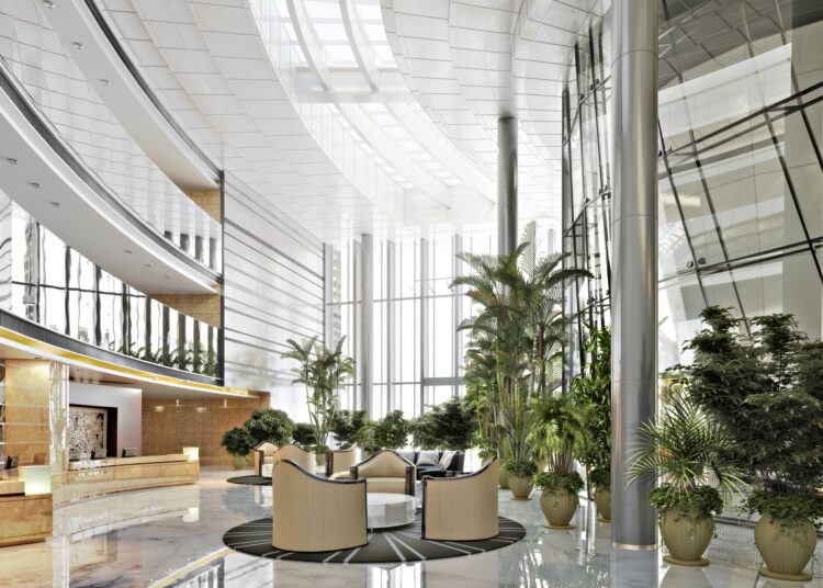 A building with a large lobby. There are ceiling-high windows in the back ground and greenery all around.