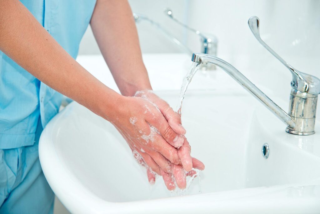 A person wearing blue scrubs washes their hands at a sink. Their hands are sudsy and pressed together under the running faucet.