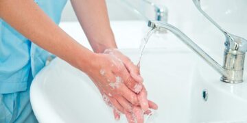 A person wearing blue scrubs washes their hands at a sink. Their hands are sudsy and pressed together under the running faucet.