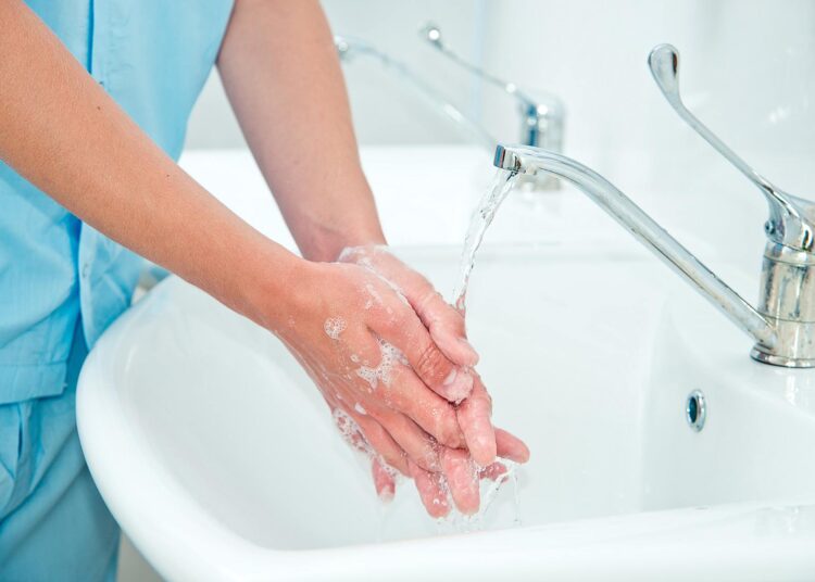 A person wearing blue scrubs washes their hands at a sink. Their hands are sudsy and pressed together under the running faucet.