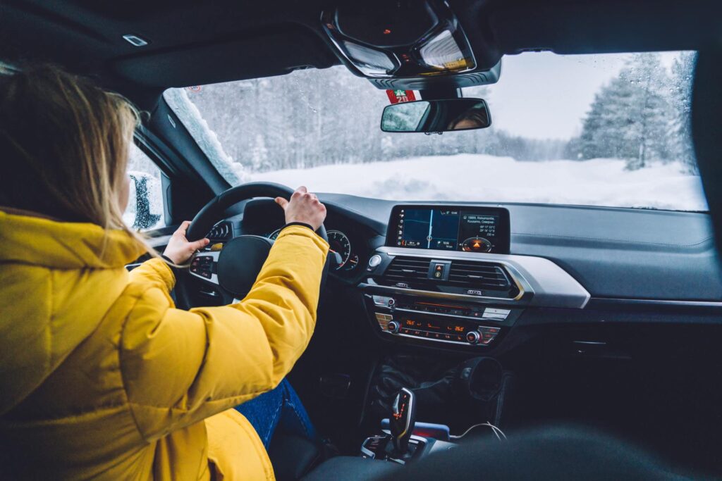 A person wearing a yellow jacket driving a car on a snowy day. The road is covered in snow and surrounded by trees.