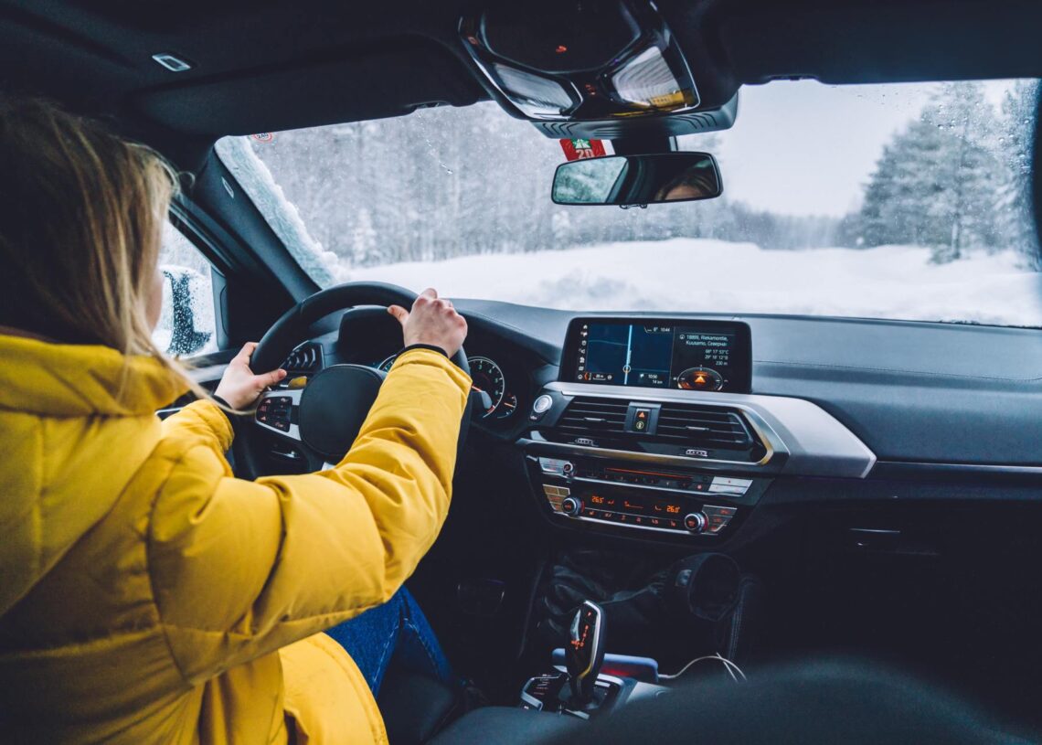A person wearing a yellow jacket driving a car on a snowy day. The road is covered in snow and surrounded by trees.