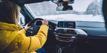 A person wearing a yellow jacket driving a car on a snowy day. The road is covered in snow and surrounded by trees.