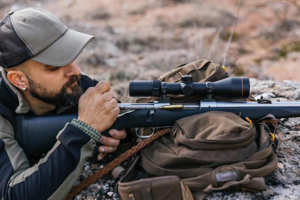 A man lays on the ground with his bolt-action rifle resting on top of his backpack. He prepares the gun for shooting.