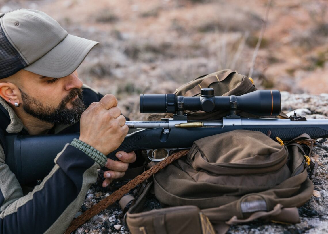 A man lays on the ground with his bolt-action rifle resting on top of his backpack. He prepares the gun for shooting.