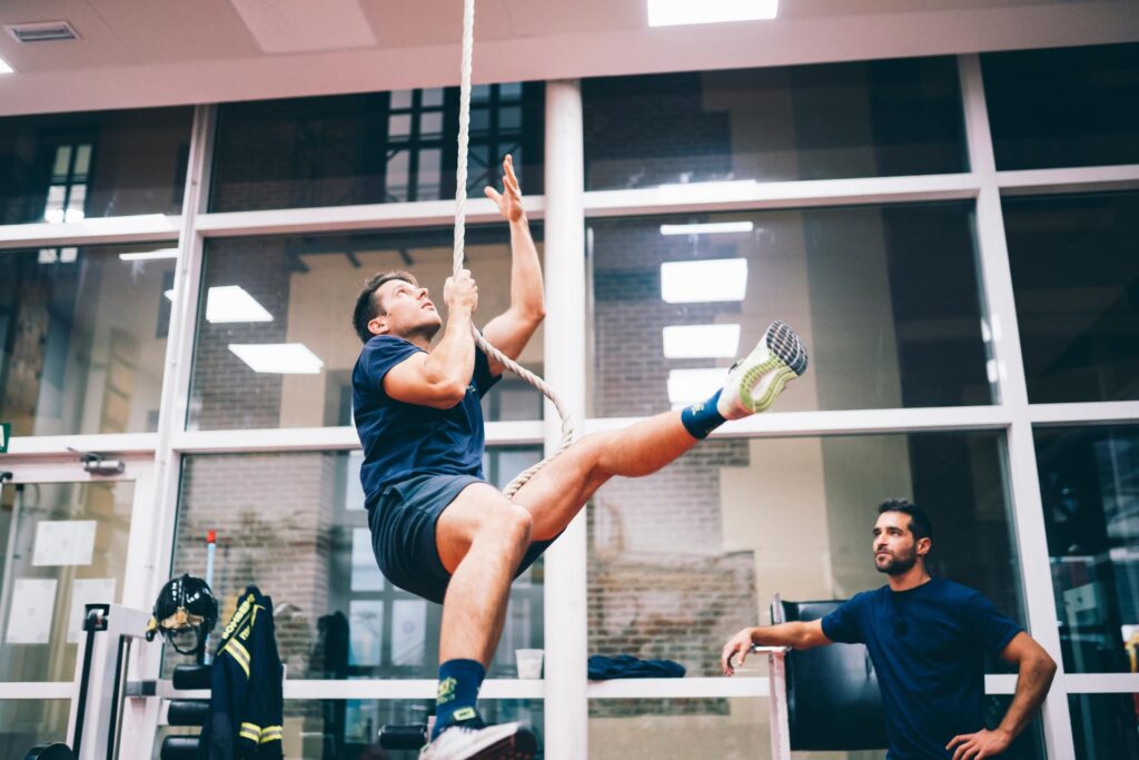 A young firefighter cadet climbs up a single rope in an indoor gym while a supervisor observes nearby.