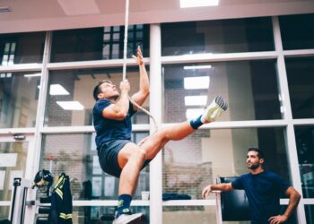 A young firefighter cadet climbs up a single rope in an indoor gym while a supervisor observes nearby.