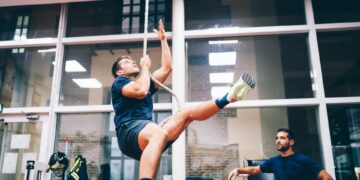 A young firefighter cadet climbs up a single rope in an indoor gym while a supervisor observes nearby.