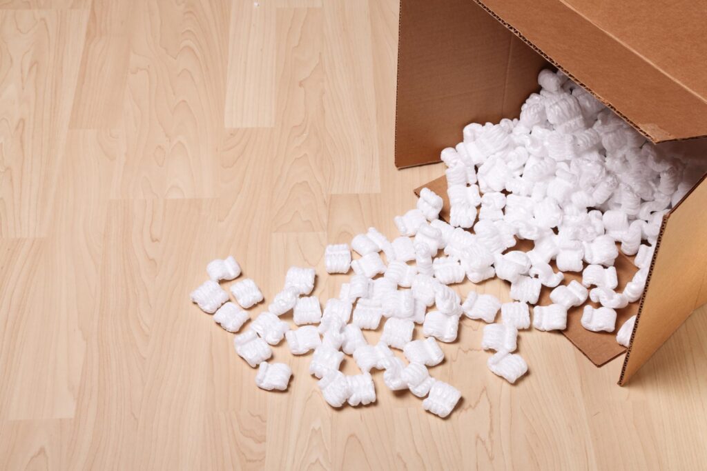 An open cardboard box filled with white packing peanuts has tipped over, spilling them onto a hardwood floor.