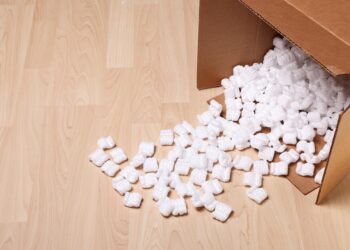 An open cardboard box filled with white packing peanuts has tipped over, spilling them onto a hardwood floor.