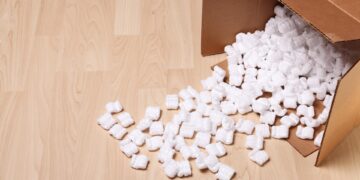 An open cardboard box filled with white packing peanuts has tipped over, spilling them onto a hardwood floor.