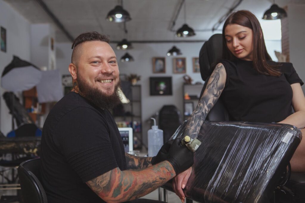 A smiling male tattoo artist in a black T-shirt and black gloves tattoos a young woman's right forearm.