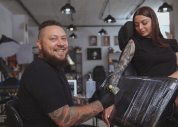 A smiling male tattoo artist in a black T-shirt and black gloves tattoos a young woman's right forearm.