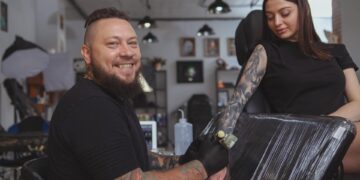 A smiling male tattoo artist in a black T-shirt and black gloves tattoos a young woman's right forearm.