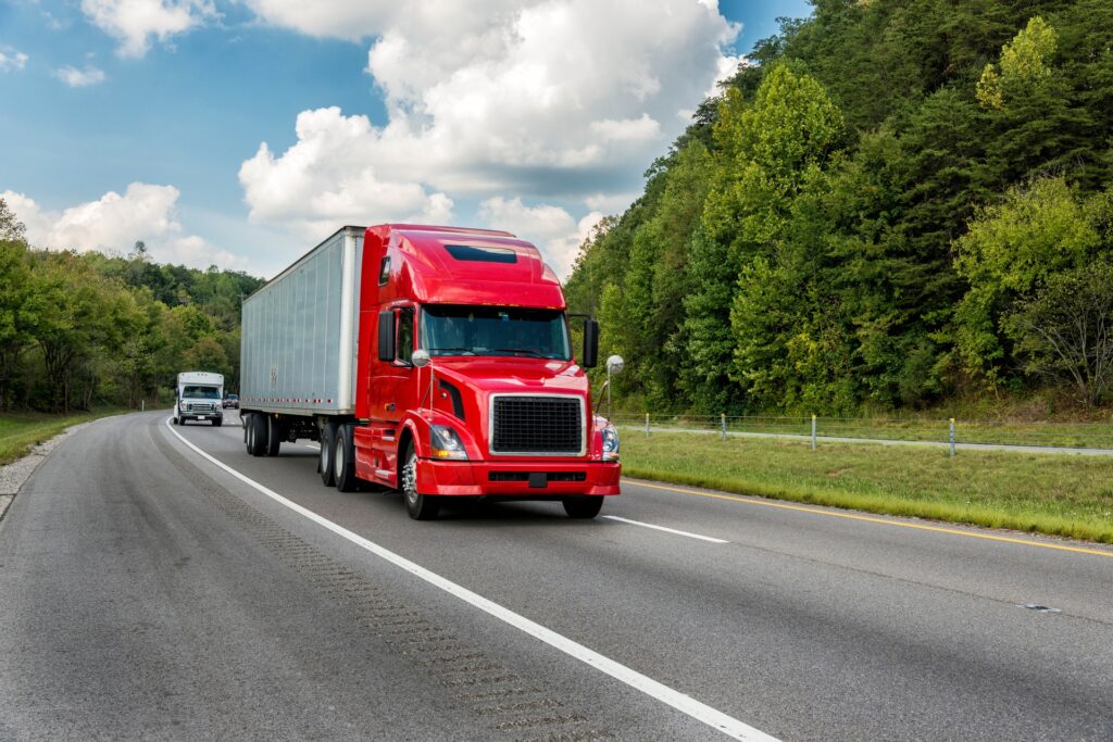 A red-cab semi-truck drives along a tree-lined two-way road on a cloudy day, with cars visible in the background.