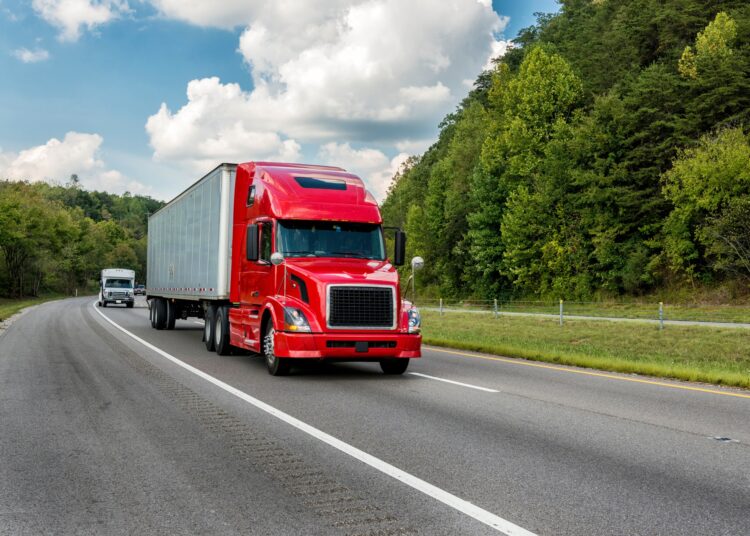 A red-cab semi-truck drives along a tree-lined two-way road on a cloudy day, with cars visible in the background.