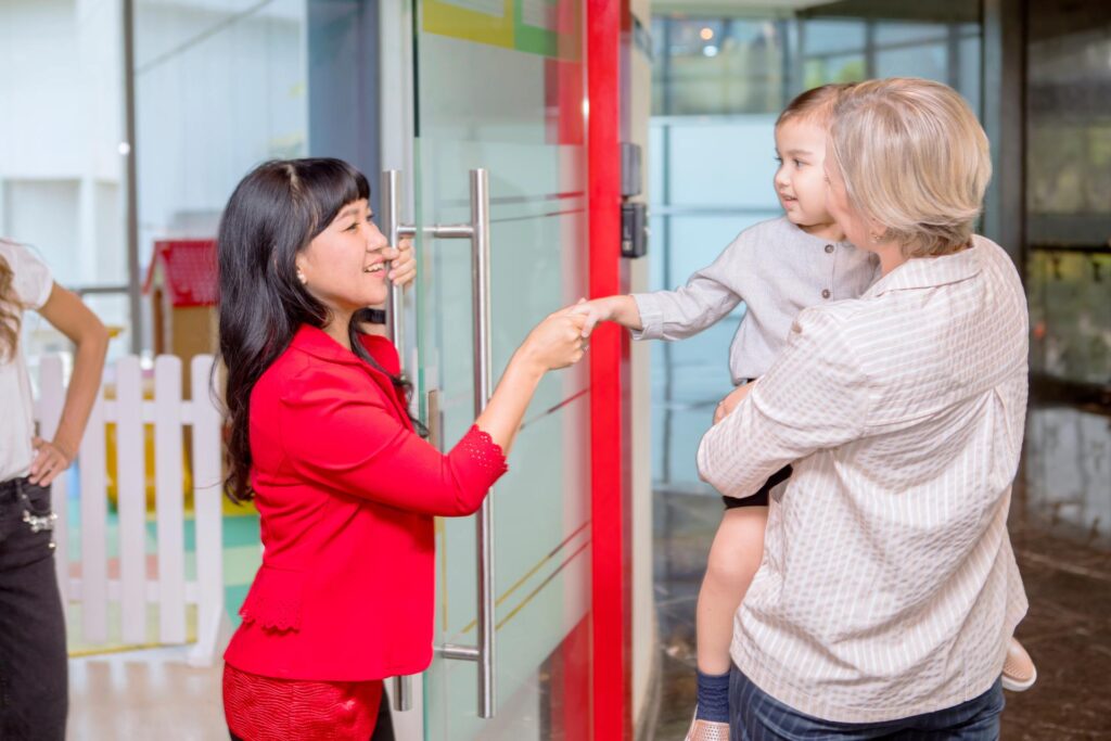 A little girl in a red dress shakes hands with her smiling teacher, her parents holding her in their arms.