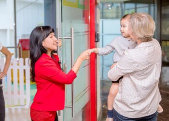A little girl in a red dress shakes hands with her smiling teacher, her parents holding her in their arms.
