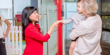 A little girl in a red dress shakes hands with her smiling teacher, her parents holding her in their arms.