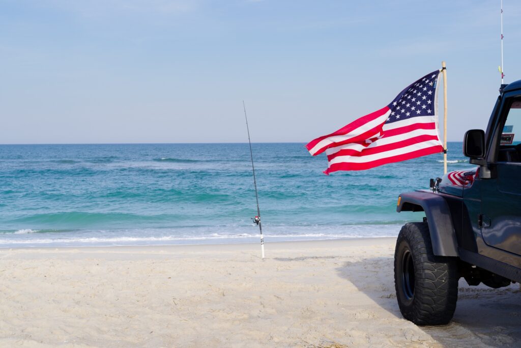 A black car parks by the beach, right next to a fishing pole. In front, an American flag sways in the wind.