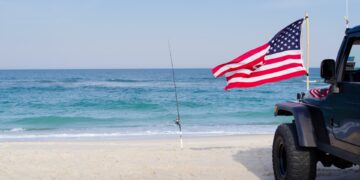 A black car parks by the beach, right next to a fishing pole. In front, an American flag sways in the wind.