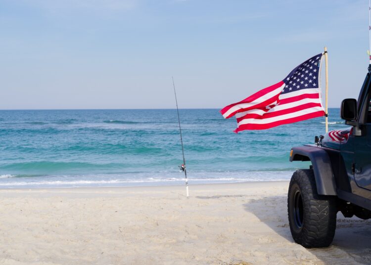 A black car parks by the beach, right next to a fishing pole. In front, an American flag sways in the wind.