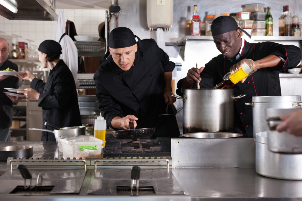 A chef wearing a black uniform, holding a pan above the stove, with multiple workers around him in a restaurant kitchen.