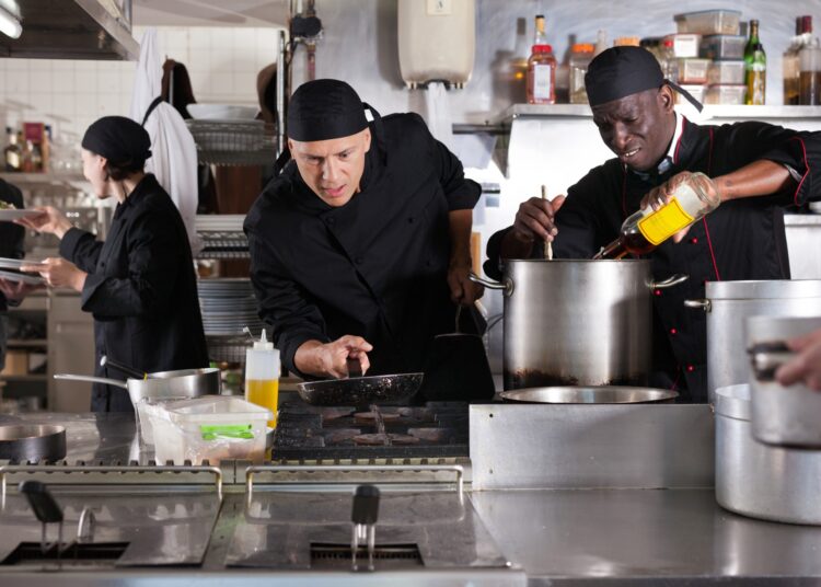 A chef wearing a black uniform, holding a pan above the stove, with multiple workers around him in a restaurant kitchen.