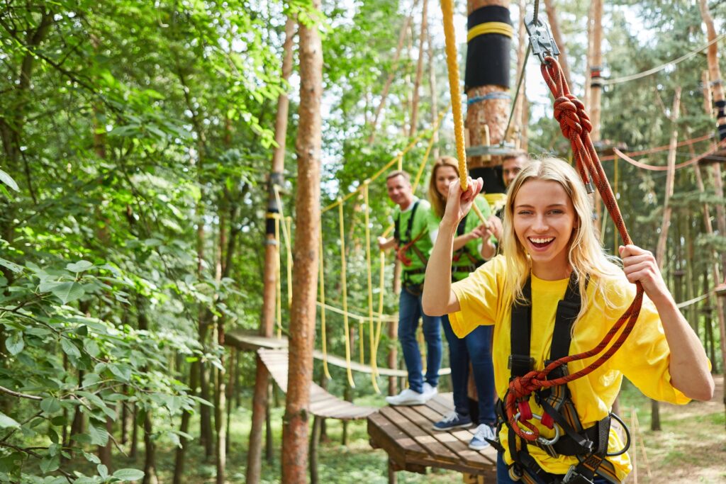 A group of people on a forest ropes course, wearing harnesses. A girl in a yellow t-shirt smiles with excitement.