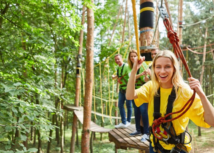 A group of people on a forest ropes course, wearing harnesses. A girl in a yellow t-shirt smiles with excitement.