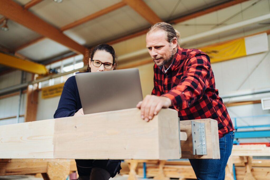 A carpenter in a red and black buffalo plaid top stands with a second carpenter at a laptop near a workstation.