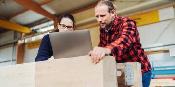 A carpenter in a red and black buffalo plaid top stands with a second carpenter at a laptop near a workstation.