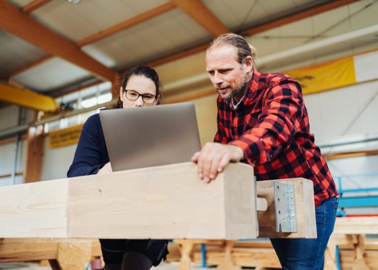 A carpenter in a red and black buffalo plaid top stands with a second carpenter at a laptop near a workstation.