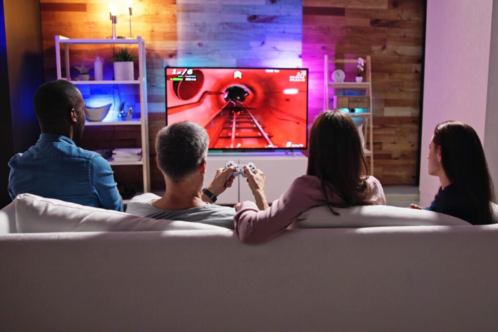 Four people on a white couch facing a large TV in a game room. Two people are playing a rollercoaster video game.