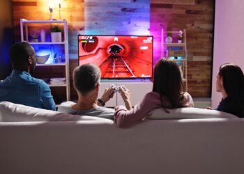 Four people on a white couch facing a large TV in a game room. Two people are playing a rollercoaster video game.