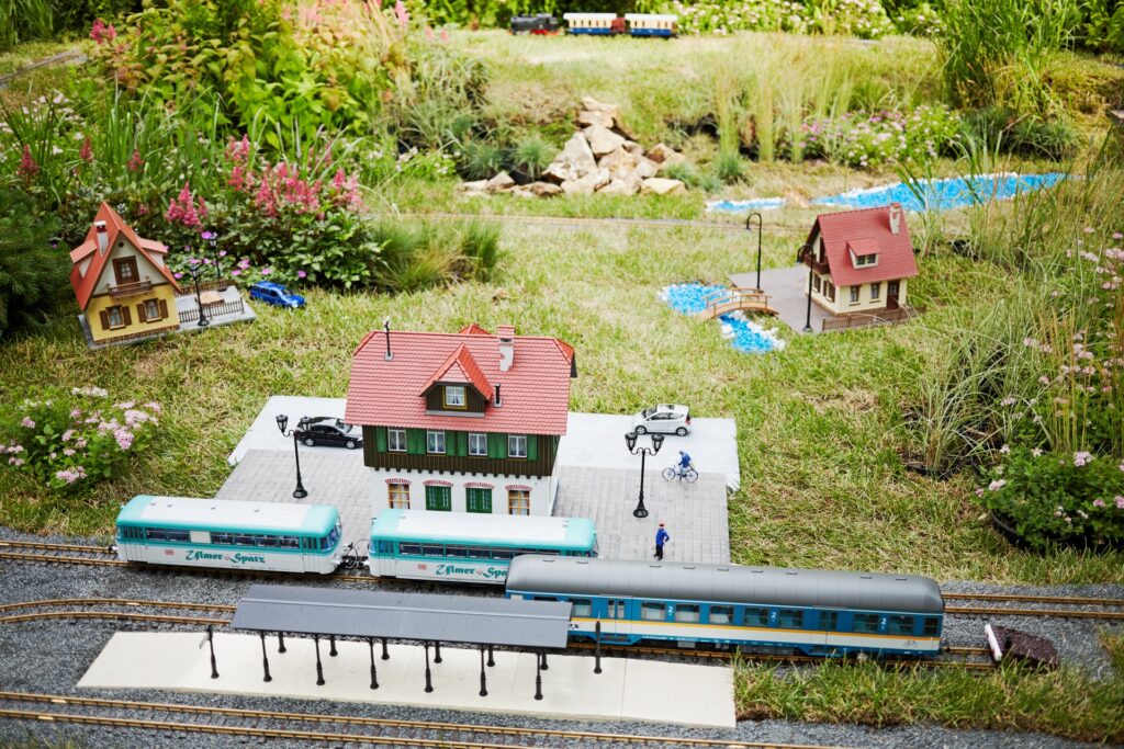 A miniature train station and tracks running through a backyard landscape. Other miniature house models rest in the grass, too.