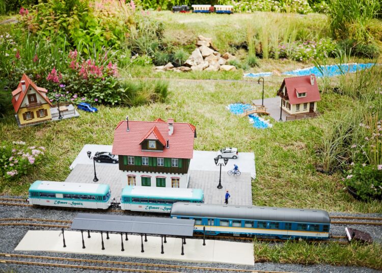 A miniature train station and tracks running through a backyard landscape. Other miniature house models rest in the grass, too.