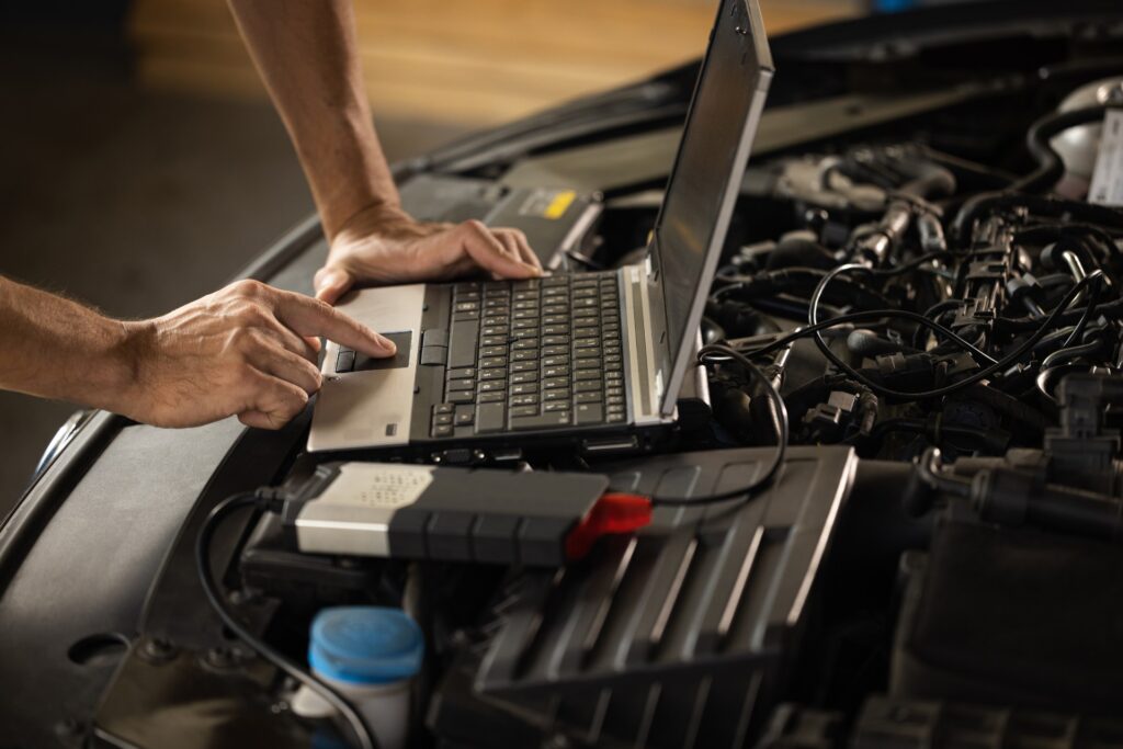 A person uses a laptop atop a car engine bay to run diagnostics with cables and tools connected to the vehicle.