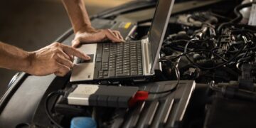 A person uses a laptop atop a car engine bay to run diagnostics with cables and tools connected to the vehicle.